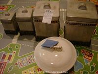 Four rectangular ceramic canisters with lids arranged alongside a round ceramic bowl with lid, all in beige glaze with decorative and metal details.