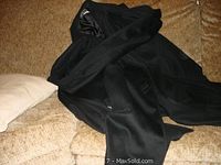 Overall view of the black wool coat laid on a couch, showing the fabric and attached scarf.
