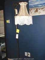Full view of the brass floor lamp showing the fabric lampshade with long white fringe hanging from the scalloped edge, mounted on a brass pole, against a dark blue wall.