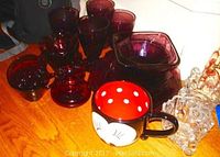 Various purple glass items including 4 glasses, 3 bowls, 5 cups and saucers, a red glass vase, and a large novelty coffee mug with cartoon face, placed on wooden surface.