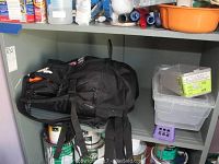 Photo of black backpack and plastic storage bins on shelf with some paint cans below, showing storage environment and condition