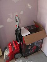 Front view of red and gray upright Rug Doctor deep carpet cleaner next to its original box. Cord is wrapped around the handle.