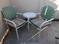 Two green woven strap patio chairs with white metal frames and striped cushions positioned around a small white metal round table on concrete surface, against beige wall.