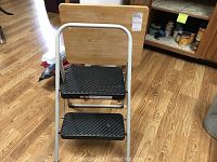 Front view of the Cosco 2-step folding step stool positioned in front of the wooden folding TV tray showing both items fully assembled and standing upright on a wood floor.