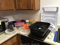 Countertop with black roasting pan, plastic containers with colorful lids, white dish rack, and kitchen utensils.