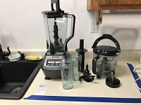 Ninja blender with large blending jar, two smaller cups, food processor bowl, multiple attachments on counter near sink.