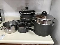 Photo showing various stacked pots, pans, and lids including a large black pot with metal steamer insert and lid, smaller pots and lids stacked on a kitchen counter.