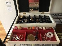 Open white jewelry box showing red velvet bottom tray with rings, bracelets and pendants; black felt upper tray partially visible