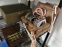 Wide view of open wood jewelry box with assorted bracelets and beaded necklaces spilling out, next to a greenish box likely a music box