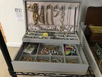 Open jewelry box showing upper lid with necklaces hanging on hooks and first tray with multiple compartments containing earrings and other jewelry pieces.