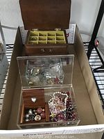 Wooden jewelry box open showing divided compartments with rings, clear acrylic unicorn box and glass box without lid containing assorted jewelry on cardboard surface.