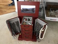 Front view of open wood jewelry box showing mirror, drawers, side panel hooks with necklaces, and upper compartment with brooches and rings.