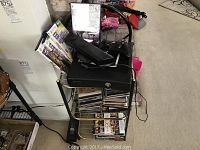 Black Xbox 360 console on top of a metal cart along with CDs and DVDs and Xbox 360 games stacked underneath and beside it.