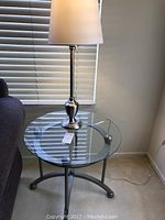 Two images of a round glass and metal side table with a metal base table lamp with white shade placed on top