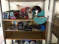 Wide shot of wooden cabinet shelves with assorted games, party items, bowling ball, and other collectibles arranged on three shelves.