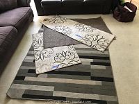 Four rugs laid out on the floor near couches, including one large area rug with gray and black stripes, two smaller beige rugs with black patterns, and one beige runner with similar black scrollwork.