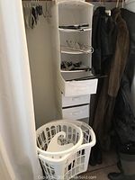 Photo showing white fabric hanging closet organizer with shelves and three fabric drawers, with coats and hangers around it, and two white plastic laundry baskets in front.