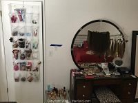 Wide view of vanity dresser with large round mirror displaying various makeup items, hair extensions hanging, and an over-the-door pocket organizer with small items.