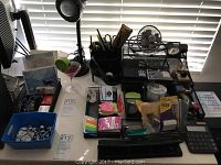 Office supplies including desktop organizers filled with pens, a small fan, tape dispensers, sticky notes, and calculator on desk near window blinds.