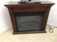 Front view of Twin Star electric fireplace with a wooden mantel and simulated burning logs visible behind glass. Remote controls placed on top.