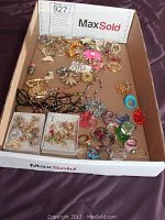 Wide layout of mixed costume jewelry, showing rings, brooches, pendants, earrings, and bracelets arranged loosely in a box.