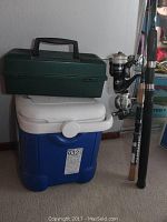 Photo showing green tackle box resting on top of a blue and white cooler, two fishing rods leaning adjacent to cooler and tackle box.