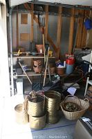 View showing Black and Decker lawnmower, brass tone planters stacked with rope handles, garden tools, and assorted pots on shelves and floor.