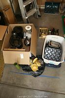 Top-down photo showing a box with a toaster, a tea kettle, a blender, a coffee maker, and a Crock Pot. Nearby are trays of flatware and stacked baking dishes and plates.