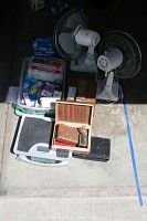 Overview of lot items including two white electric fans, plastic bins containing health supplies, digital scale, carved boxes, and Wahl shaver accessories spread on ground.