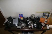 Photo showing multiple electronics including Amazon e-readers, Polaroid 600 camera with film, Ultronic camera, vintage red radio, and assorted cords and chargers laid out on a table