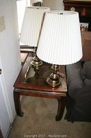 Photo showing one end table with clear glass top, wooden frame, and two brass lamps with white pleated shades on top next to a wall and couch.