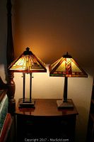 Two Tiffany style table lamps illuminated on a wooden side table next to a bedpost, displaying warm amber and earth tone stained glass shades with geometric designs.