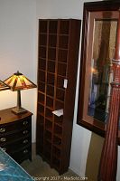 Photo of one dark brown tall wooden CD shelf with many compartments, placed in a room near a side table and lamp.
