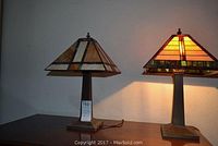 Two Tiffany-style table lamps side by side on a wooden surface, both lamps turned on. The left lamp shade features cream and amber stained glass with multi-colored geometric accents; the right lamp shade has yellow and amber glass with green accents.