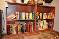 Two wooden bookcases filled with various paperback and hardcover books arranged on shelves, with some decorative items such as seashell and small basket hat visible on top.