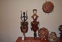 Photo showing two large Guro masks, two circular carved wooden pieces with cowrie shells, and smaller carved sculptures on display table against wall.