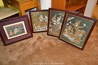 Four framed pieces of Asian wall art arranged on carpeted floor: three are silk fabric artworks, the fourth is a painting of Krishna.