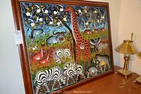 Front view of large colorful framed painting showing African animals including zebras, giraffe, elephant, rhinoceros, lions, and various birds. Two brass lamps on table beside painting.