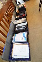 Eight bins arranged in a row along hallway. Visible are various binders, paper packages, notepads, and office supplies inside the bins.