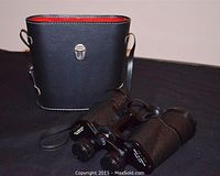 Binoculars placed in front of the black leather carrying case with red interior lining visible, showcasing the clasp and strap.