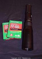 Two unopened boxes of Fujifilm FP-100C instant color film and the Opticam 500mm lens placed on a black surface.