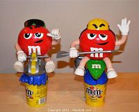 Photo shows 2 red M&M's candy dispensers, one with black cap holding a saw, other with yellow hat holding a spear. Both mounted on yellow candy containers.