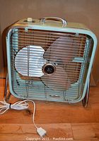 Front view of the Electrohome Long Life box fan showing three silver metal blades, metal protective grille, and electrical cord with plug.