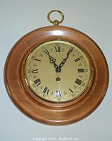 Front view of the round wooden clock with golden face, black Roman numerals, and brass-framed glass cover closed.