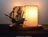 Desk lamp featuring a miniature 'Santa Maria' sailing ship model on an oval teak base with a rectangular cream-colored textured lampshade emitting warm light