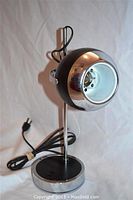 Full view of desk lamp showing black and chrome eyeball shade, chrome support rod and circular base with power cord.