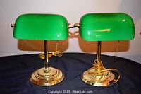 Pair of two banker's lamps with clear green glass shades and shiny gold-colored metal bases and stems. Electrical cords and pull-chains are visible indicating functionality.
