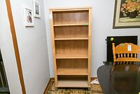 Front view of wood laminate 5 shelf bookcase placed against a wall near framed pictures and dining furniture.