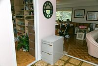 Room photo showing gray two drawer filing cabinet under the stairwell, with clock style round black framed mirror hanging on wall.
