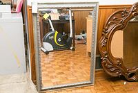 Full view of rectangular mirror with silver ornate carved frame, leaning against wall reflecting exercise bike and room interior.